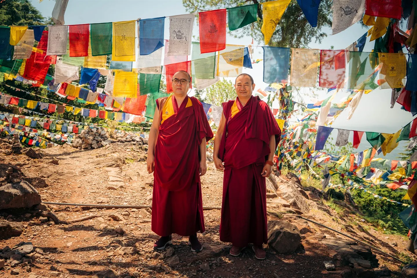Two Buddhist monks in red robes under prayer flags on a forest path